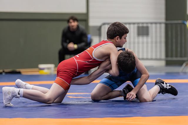 Two youth wrestlers compete intensely on a blue mat, one in red dominating from above as a coach watches from behind.