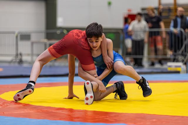 Two young wrestlers battle intensely on a yellow mat, one in red lifting his opponent in blue during a takedown attempt.