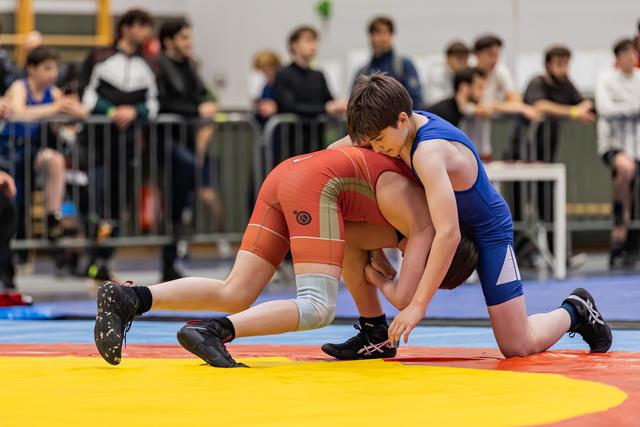 Two young wrestlers in red and blue singlets locked in a ground grapple, intense focus during an indoor tournament.