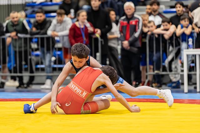 Boy in blue singlet controls opponent Yusupov M in red on yellow mat, crowd watching intently in background.