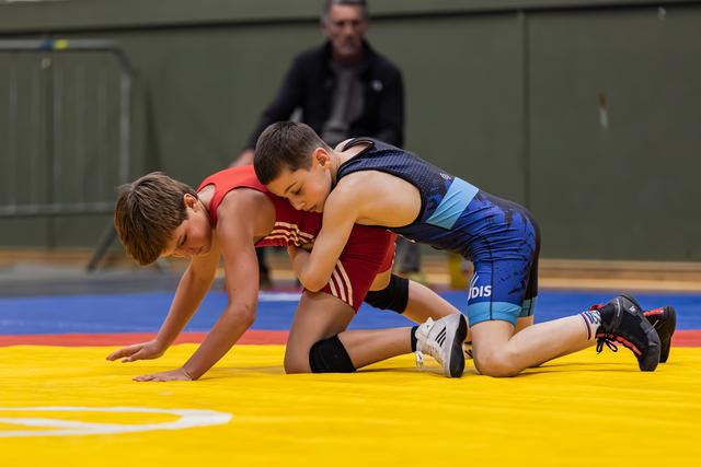 Two young boys wrestle on a yellow mat, one in blue pinning down opponent in red during indoor youth bout.