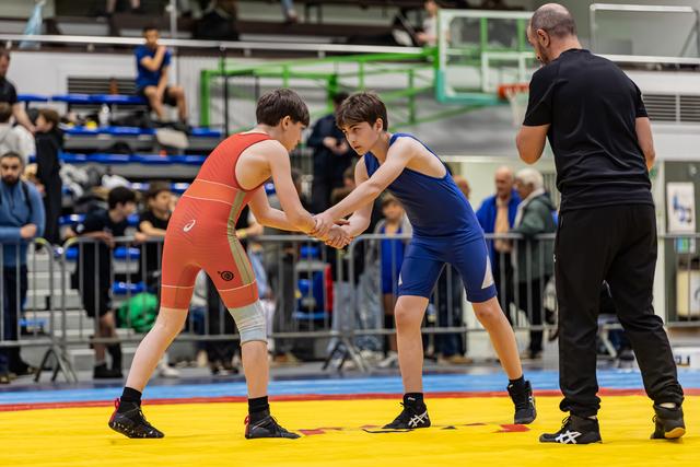 Two young wrestlers in red and blue singlets shake hands before their bout, supervised by a referee in a packed gym.