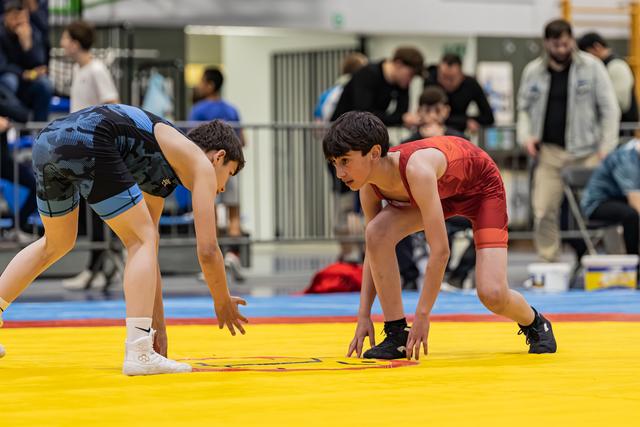 Two young wrestlers in blue and red singlets crouch in a tense face-off stance on a yellow competition mat.