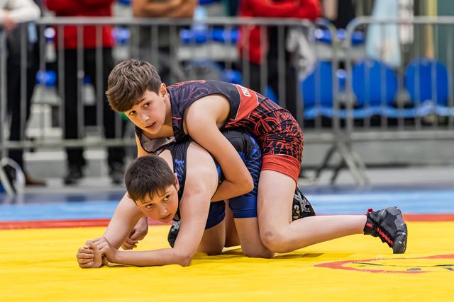 Young wrestler in black-red singlet controls opponent on yellow mat, both focused during intense bout.
