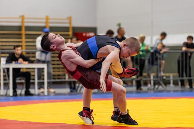 Young wrestler in red singlet hoists opponent off the mat during an intense match in a gymnasium.