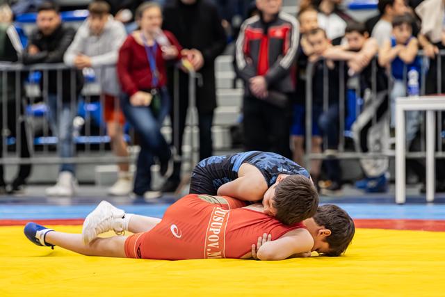 Wrestler in red singlet pinned to the mat by blue opponent, crowd watching intently in the background.