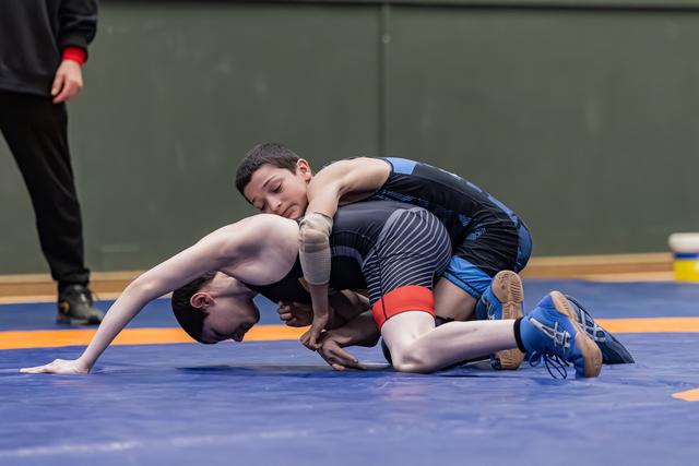 Young wrestler in black singlet controls opponent on blue mat, applying pressure with focused intensity during bout.