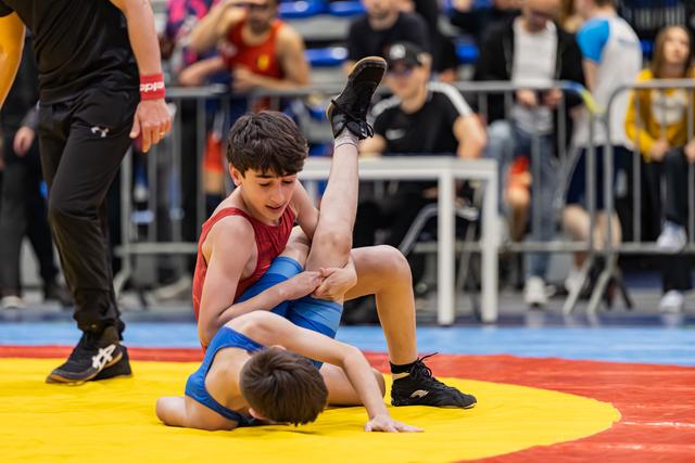 Young wrestler in red singlet grips opponent's leg on yellow mat, focused expression, referee watching nearby.