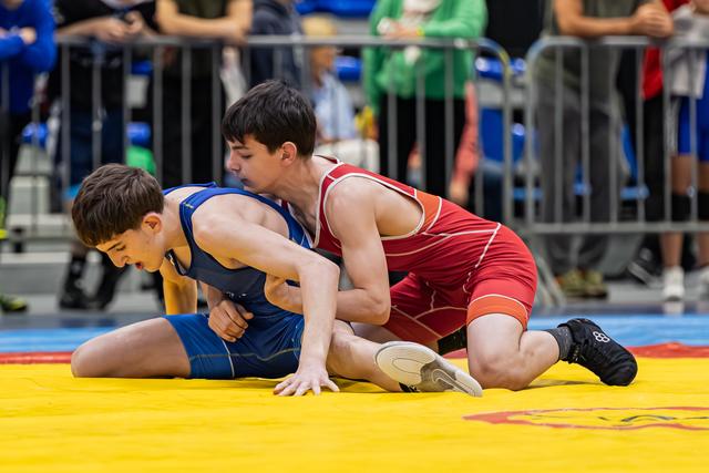 Teen in red singlet pins opponent in blue to yellow mat, both showing intense focus during match.