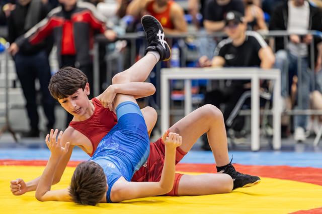 Young wrestler in red singlet dominates opponent in blue, lifting his leg on a colorful competition mat.