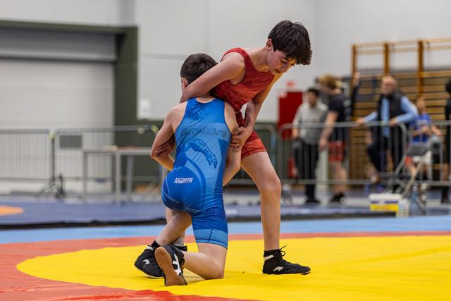 Young wrestler in red singlet dominates opponent in blue, who is down on one knee on a yellow and red mat.