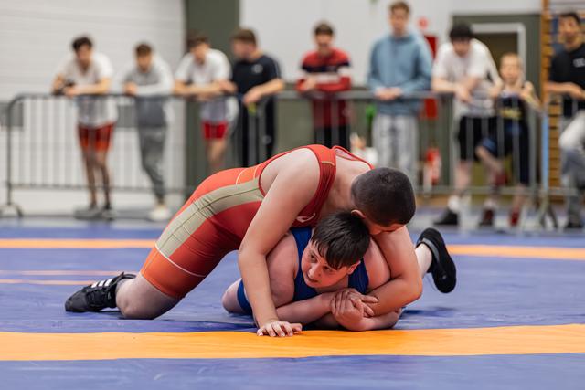 Young wrestler in red singlet pins opponent in blue to the mat while spectators watch from behind barriers.