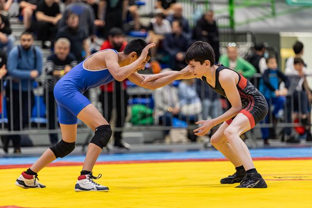Two young wrestlers in blue and black singlets square off in a tense stance, crowd watching in the background.