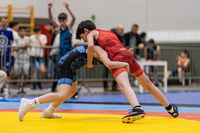 Two young wrestlers locked in a tight clinch on a yellow mat, crowd cheering energetically in the background.