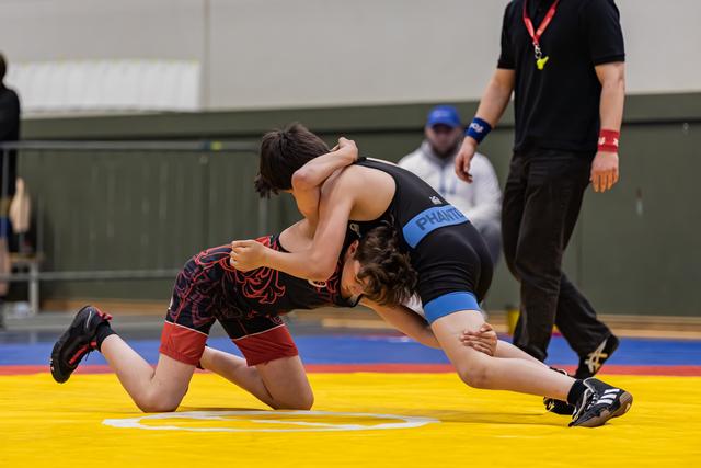 Two young wrestlers locked in a headlock battle on a yellow mat, referee observing closely in the background.