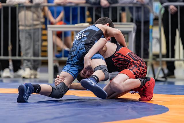 Two young wrestlers compete intensely on a blue mat, one in blue dominating from above, the other in red defending below.