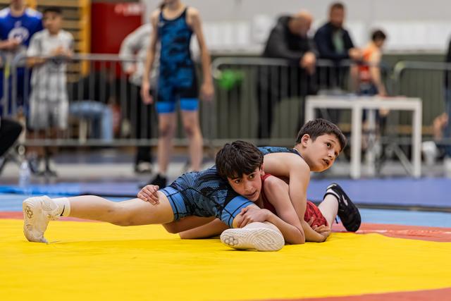 Two young wrestlers on a yellow mat, one in blue controlling his opponent from behind, both focused and determined.