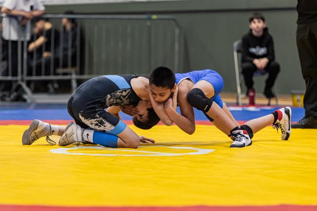 Two young wrestlers in blue and black singlets locked in a ground clinch on a yellow competition mat, focused intensity.