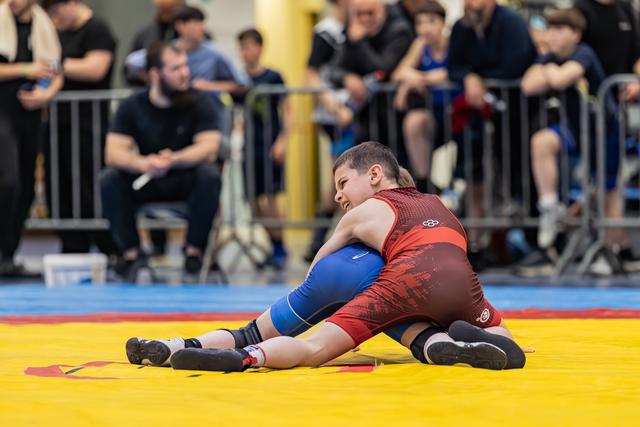 Two young wrestlers battle on a yellow mat, one in red pinning the other in blue, faces tense with effort.