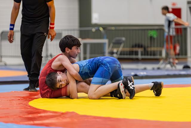 Boy in blue singlet pins opponent in red to the yellow mat while referee watches nearby in gym.