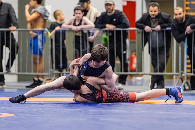 Two young wrestlers battle on the mat, one pinning the other down, with coaches and spectators watching intently behind barriers.