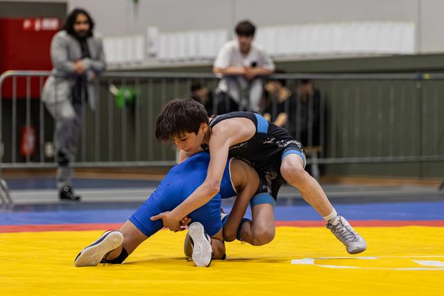 Two young wrestlers battle for control on a yellow mat, one in blue executing a takedown while spectators watch.