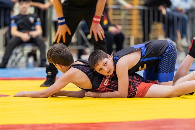 Two young wrestlers grapple on a yellow mat, one pinning the other down, face showing fierce concentration.