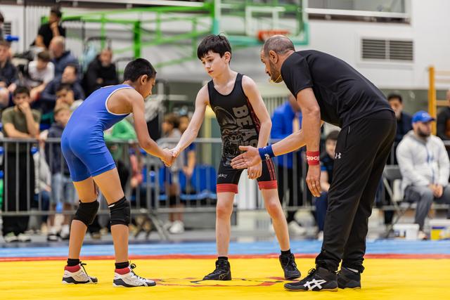 Two young wrestlers shake hands before a match as an adult referee guides them on a yellow mat in a busy gymnasium.