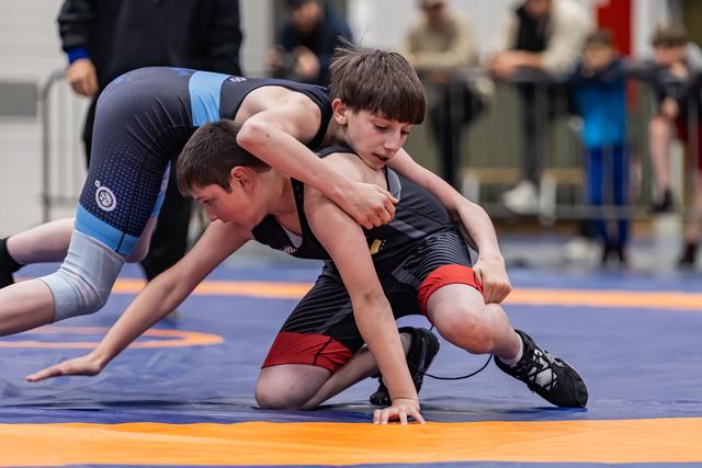 Two young boys wrestle intensely on a blue and orange mat, one in black dominating with focused determination.