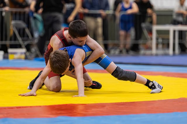 Two young wrestlers grapple on a yellow mat, one in blue controlling the other in red from above.
