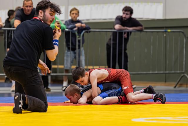 Young wrestler in red singlet pins opponent on mat while referee kneels close, watching intently in indoor sports hall.