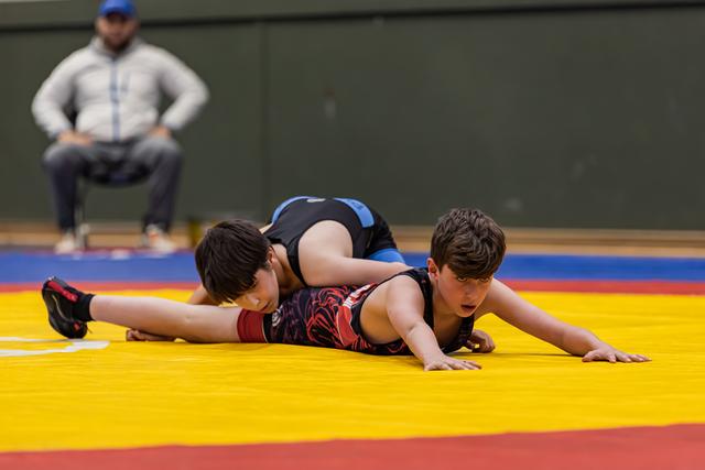 Two young boys wrestling on a yellow mat, one pinning the other down, coach watching intently in the background.