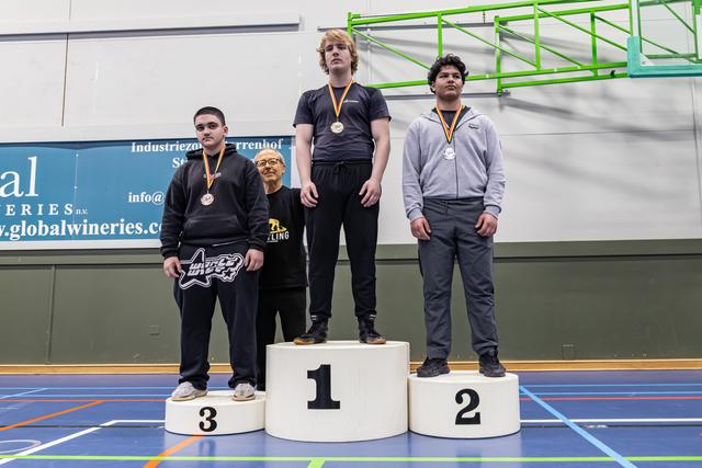 Three teenage wrestlers stand on podiums 1-3, wearing medals, with a coach beside them in an indoor sports hall.