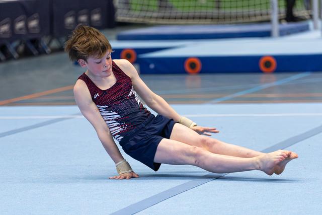 Young athlete performs seated floor routine with extended legs on blue mat at indoor gymnastics facility