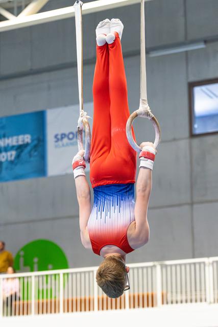 Athlete performing inverted hang on still rings with extended legs and pointed toes during indoor competition