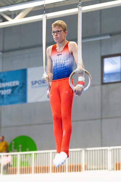 Young athlete with glasses performing still rings routine, displaying intense focus and controlled strength during apparatus work