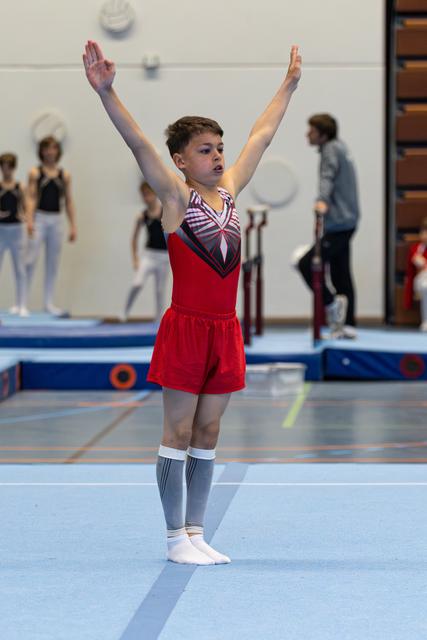 Young gymnast in red leotard completes routine with arms raised overhead, displaying focus and determination on floor mat