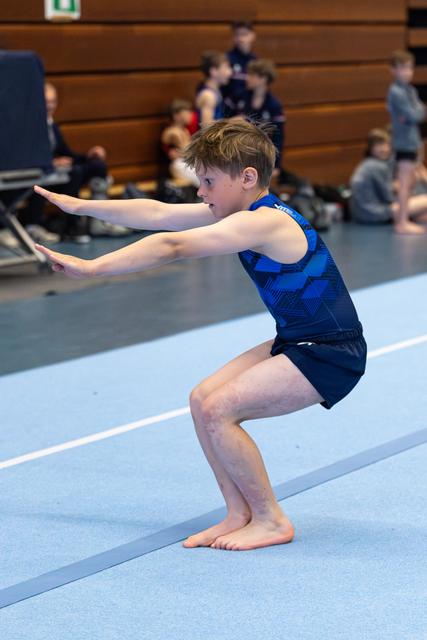 Young athlete performs floor exercise in blue leotard, focused expression in deep squat with arms extended forward