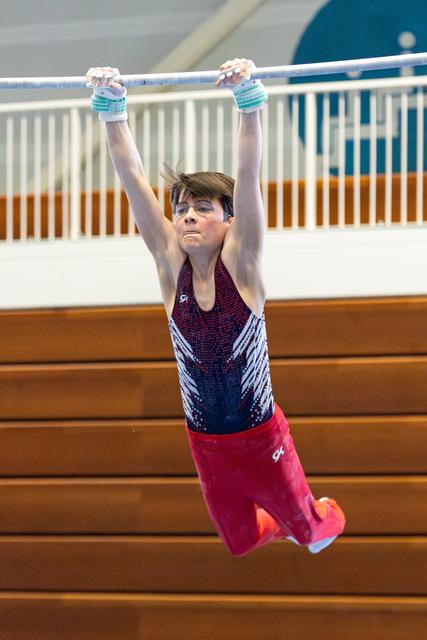 Young athlete hanging from horizontal bar with intense concentration, wearing purple leotard and red shorts in gymnasium