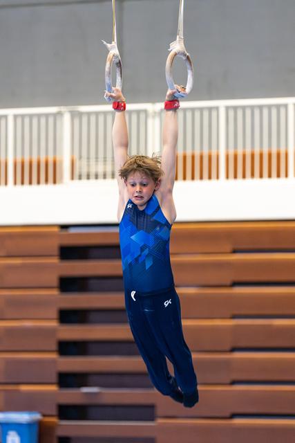 Young athlete hanging from rings with focused expression during training, wooden bleachers visible in background