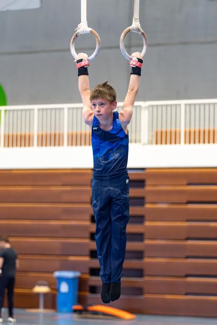 Young athlete in blue tank top hanging from still rings with focused expression during training at indoor gymnasium