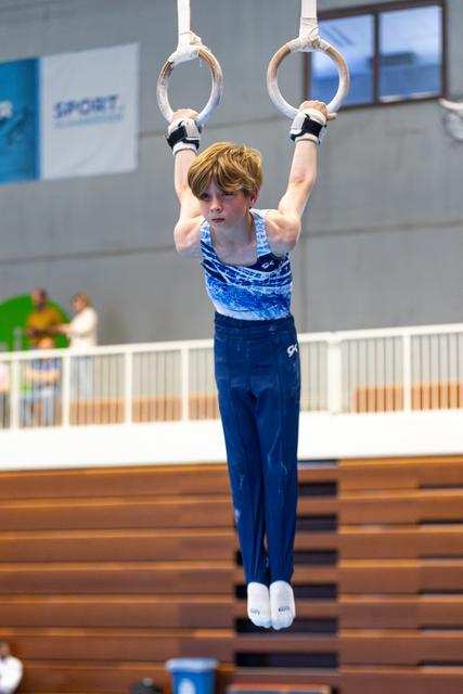Young gymnast hangs from still rings during competition, showing strength and concentration in a sports hall