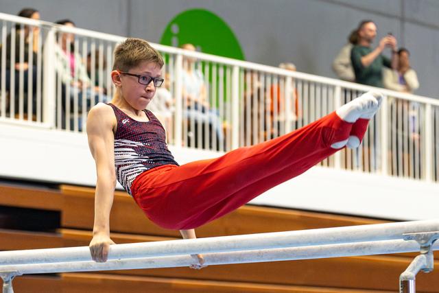 Young gymnast with glasses performs L-sit position on parallel bars while spectators watch from viewing area above