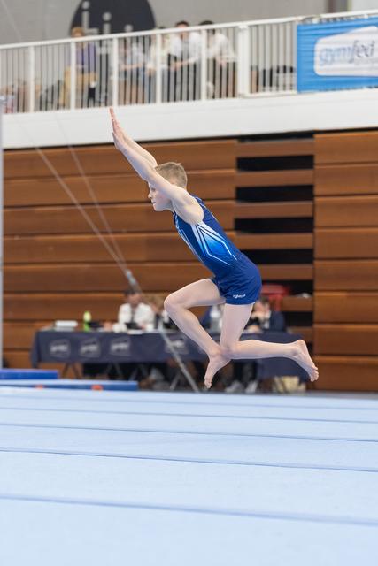 Gymnast in blue leotard performs a dynamic leap with extended arms during floor exercise routine in indoor gymnasium