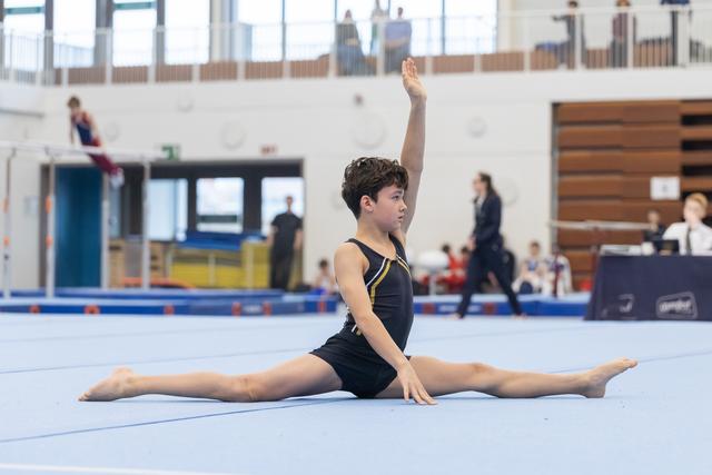 Young gymnast in black leotard performs a full split with one arm raised, demonstrating flexibility on the blue competition floor
