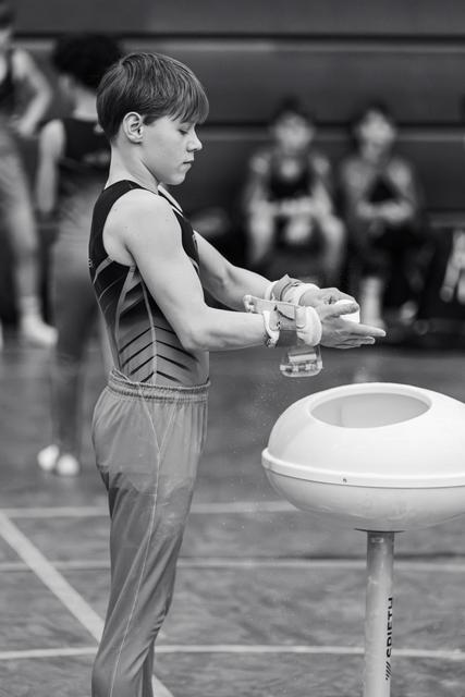 Young male gymnast chalks his hands before pommel horse routine, focused and preparing in gym with blurred figures behind