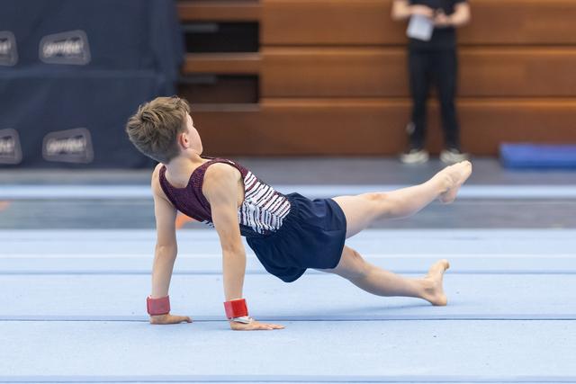 Young gymnast performs a plank-based floor exercise position with one leg extended, wearing navy and white leotard on blue mat