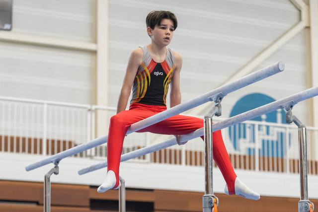 Young gymnast in colorful leotard sits on parallel bars with focused expression during training session
