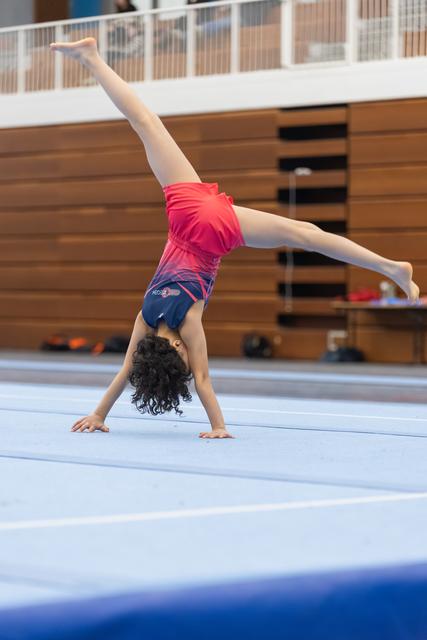 Young gymnast performing handstand on blue floor mat, pink leotard, curly hair, training in gymnasium
