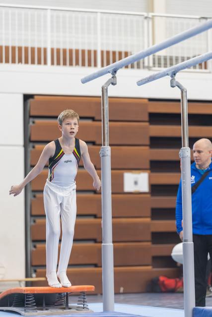 Young male gymnast lands on orange springboard with arms outstretched, coach in blue observing nearby in gymnasium